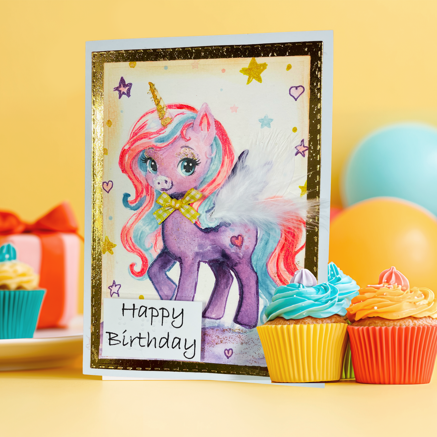 A birthday card featuring a colorful watercolor unicorn with pink, blue, and purple hair, a gold horn, and a white wing, with the words 'Happy Birthday' written on a small sign. The card is styled on a yellow surface with cupcakes and balloons in the background.