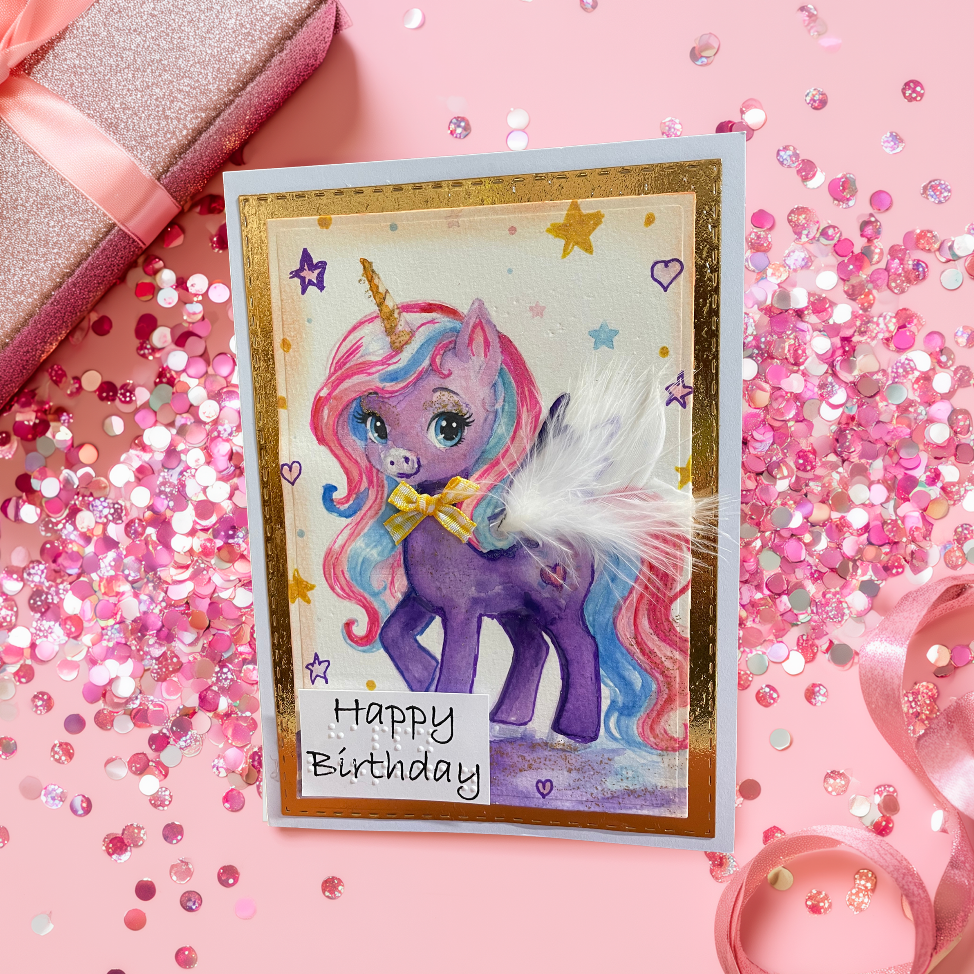 A handmade unicorn birthday card with a purple pony, pink and blue hair, and a white feather, styled on a pink background with glitter and a gift box.