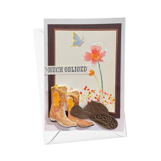 A handmade greeting card with a white envelope, featuring a western theme with brown cowboy boots, a pink flower, a blue butterfly, and the text 'MUCH OBLIGED'.