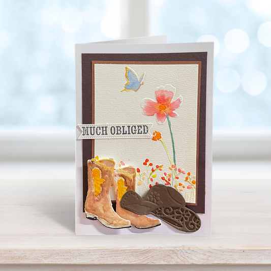 A handmade greeting card with a western theme, featuring embossed cowboy boots, a pink flower, a blue butterfly, and the text 'MUCH OBLIGED' on a light wood surface.