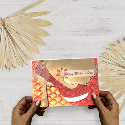 Card with high-heeled shoe design and 'Happy Mother's Day' text held by hands on a light wooden surface with decorative leaves.
