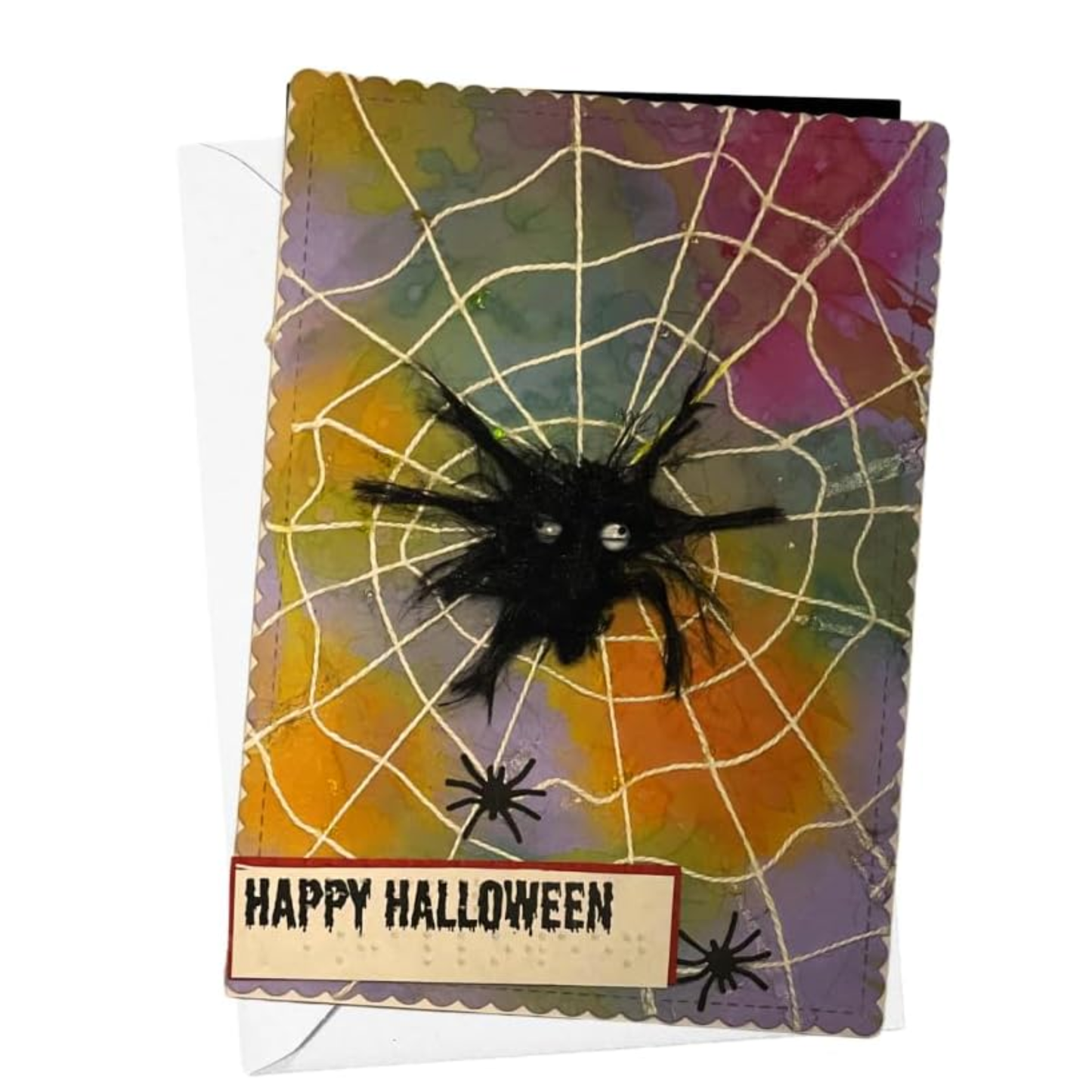 A handmade Halloween greeting card featuring a black spider on a web pattern over a multicolor watercolor background of orange, yellow, purple, and green, with a 'Happy Halloween' banner at the bottom.