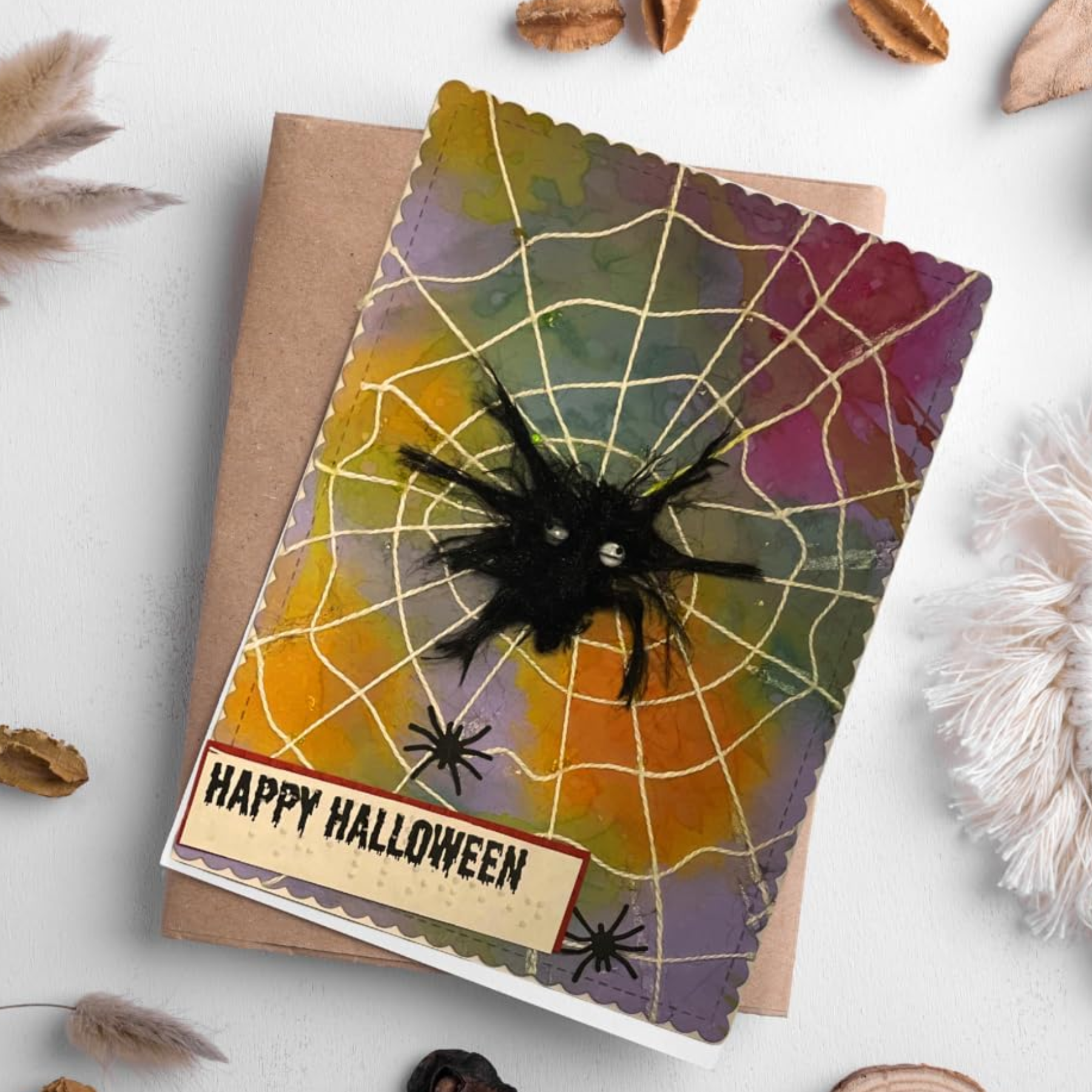 A Halloween greeting card with a spiderweb design, a black spider decoration, and the words 'HAPPY HALLOWEEN' on a kraft envelope, styled on a light surface with dried leaves and fluffy white petals.