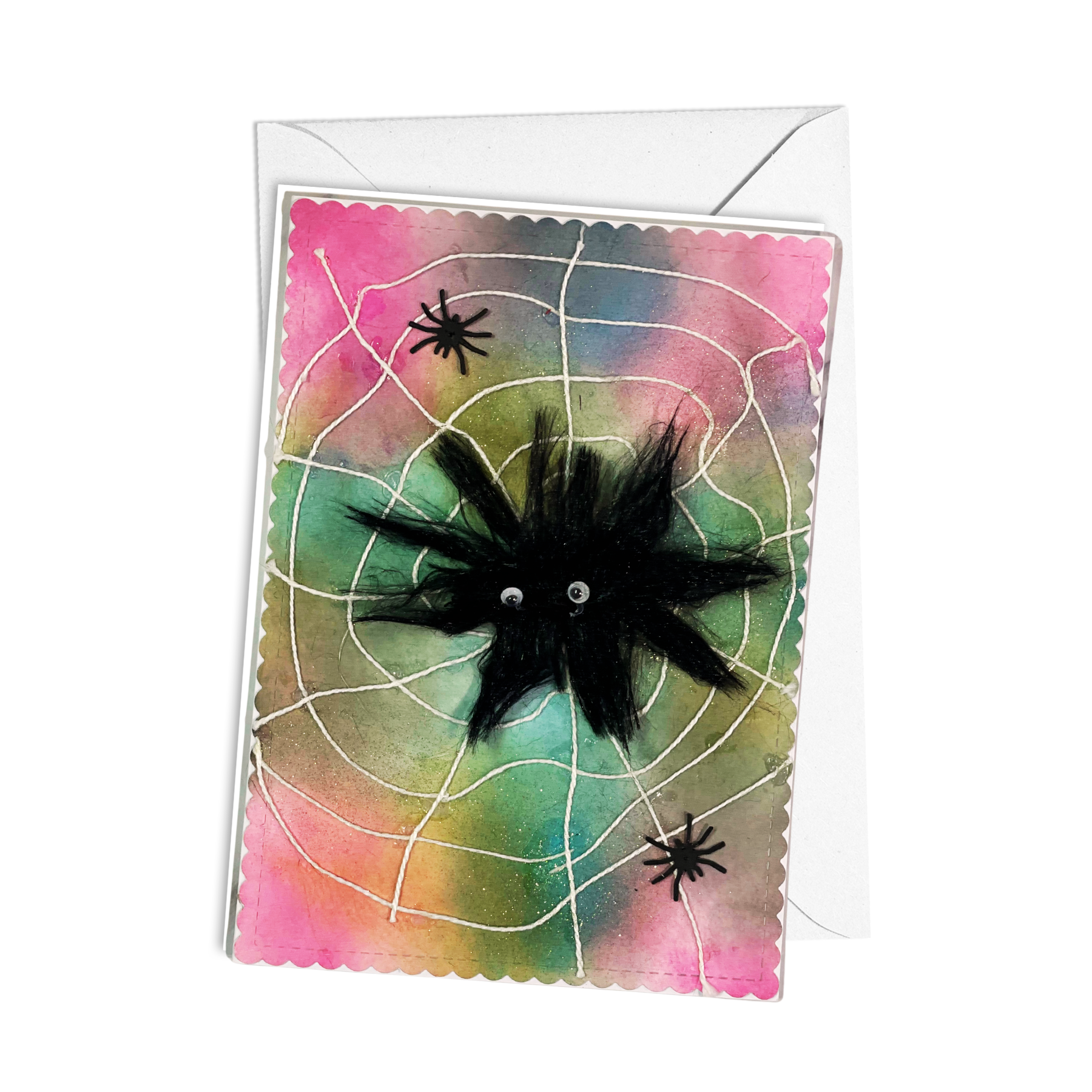 A Halloween greeting card with a white envelope, featuring a black spider on a web over a pink, green, and blue watercolor background.