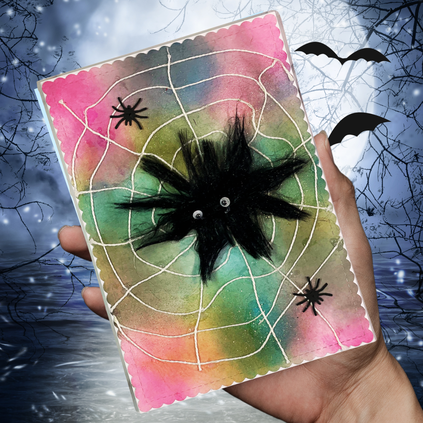 A hand holds a handmade Halloween card featuring a black fuzzy spider, white spiderwebs, and a pink, green, and blue watercolor background, set against a spooky night sky backdrop with bats and bare branches.