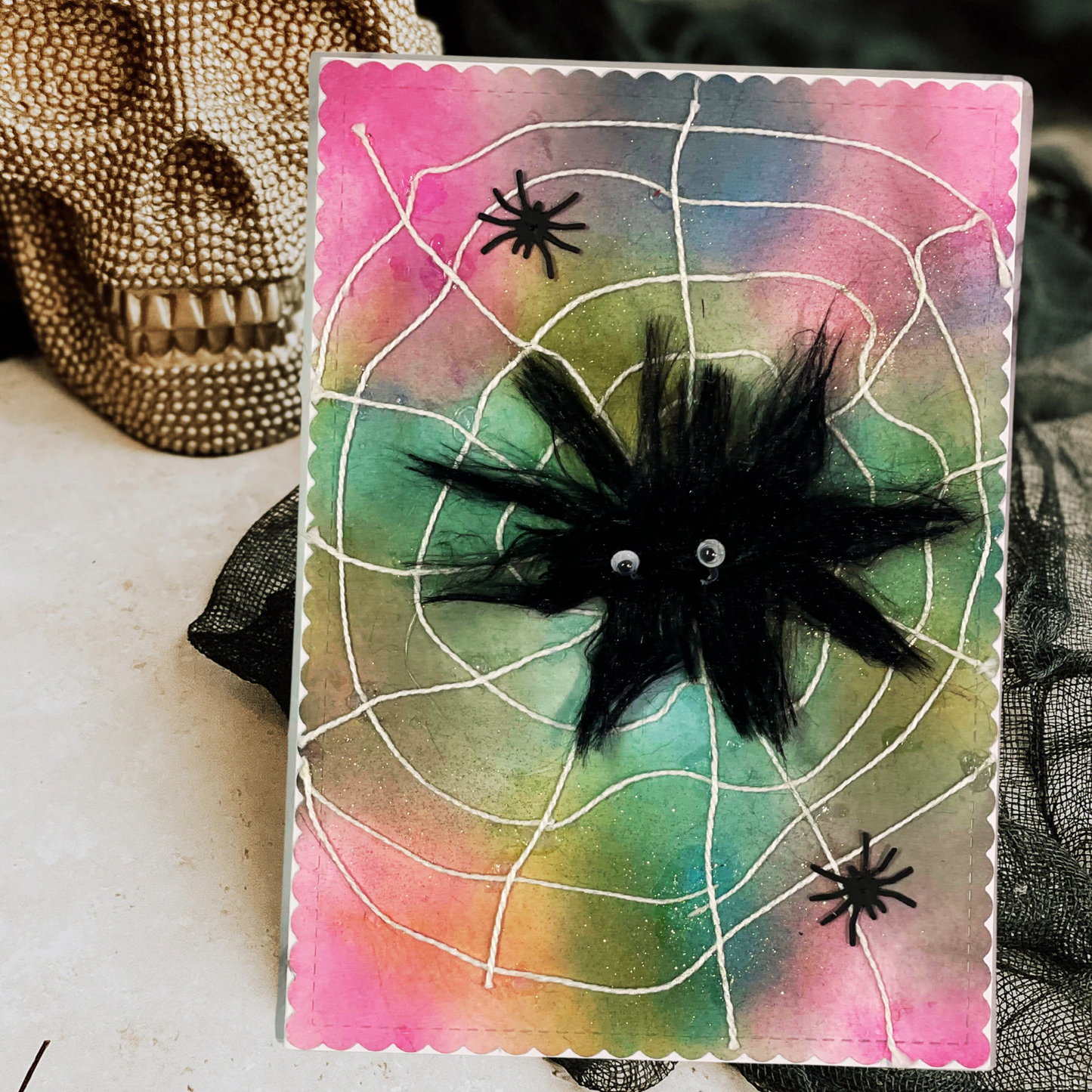 A handmade Halloween greeting card featuring a black feather spider on a pink, green, and blue watercolor background with white spiderweb lines. The card is displayed next to a decorative skull.
