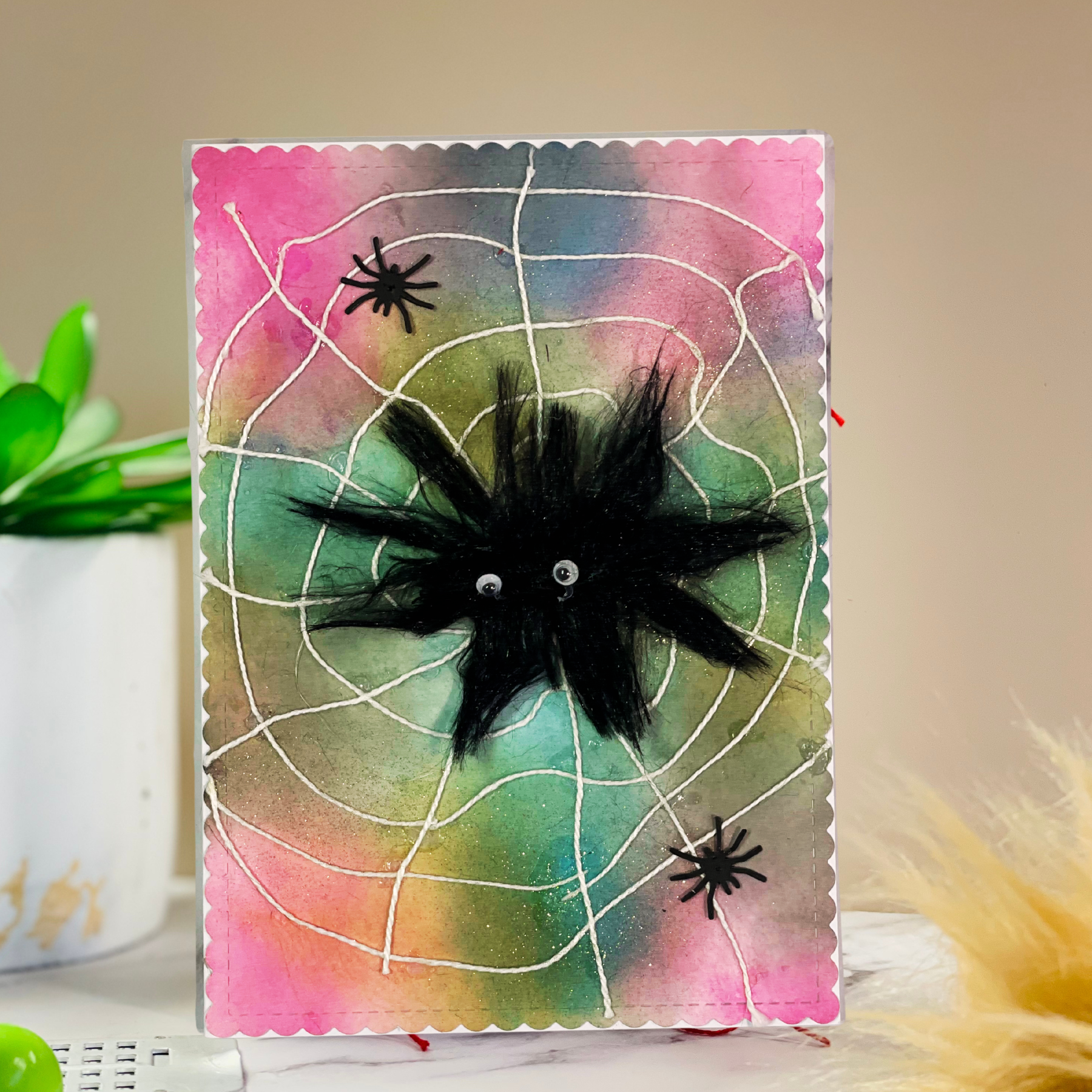 A handmade Halloween card featuring a black fuzzy spider on a colorful pink, green, and blue watercolor background with white spiderweb lines, displayed on a tabletop.