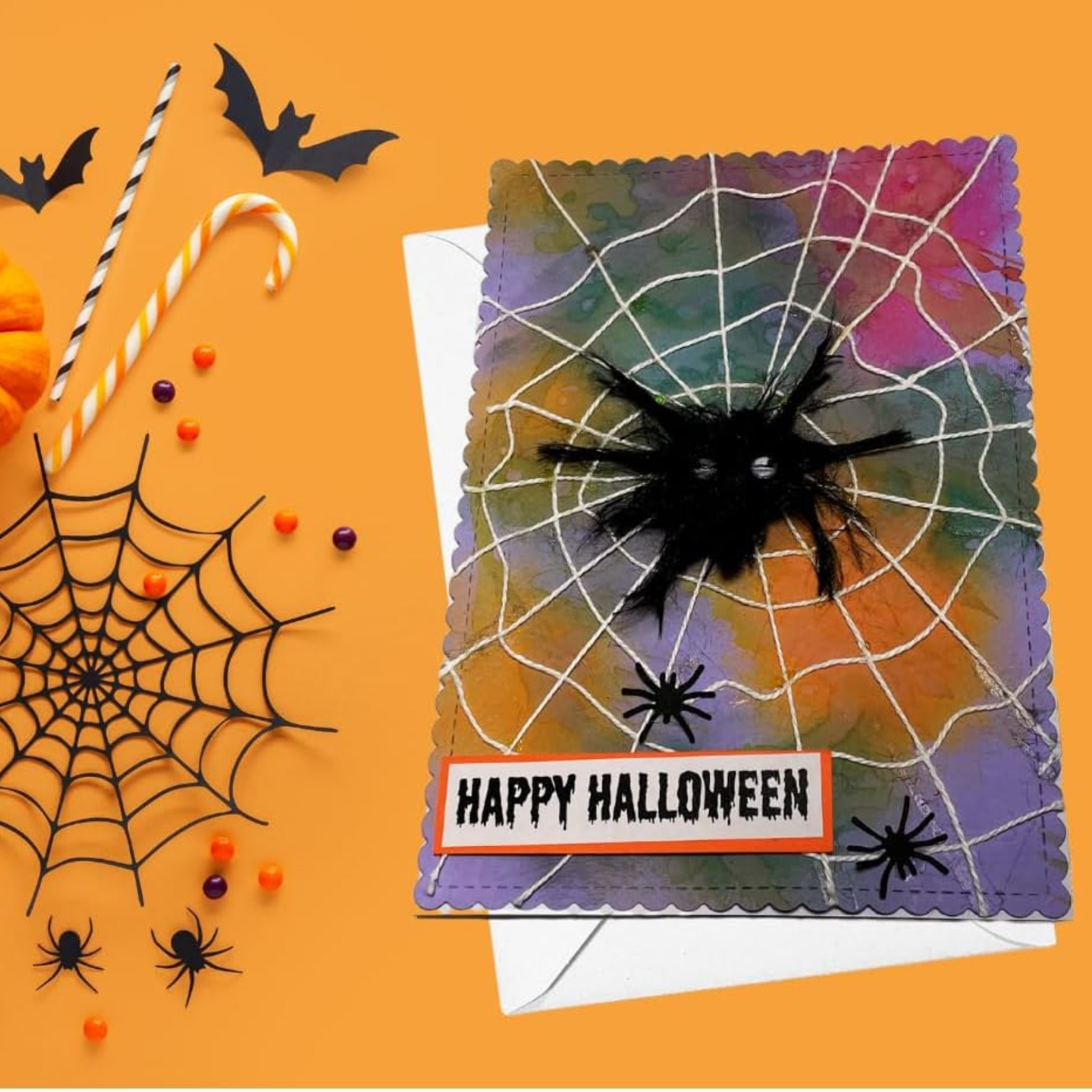 A Halloween greeting card with a colorful marbled background, white spiderweb pattern, black spider decoration, and a 'HAPPY HALLOWEEN' banner, styled on an orange surface with spiderweb cutout, candy, bats, and small orange decorations.