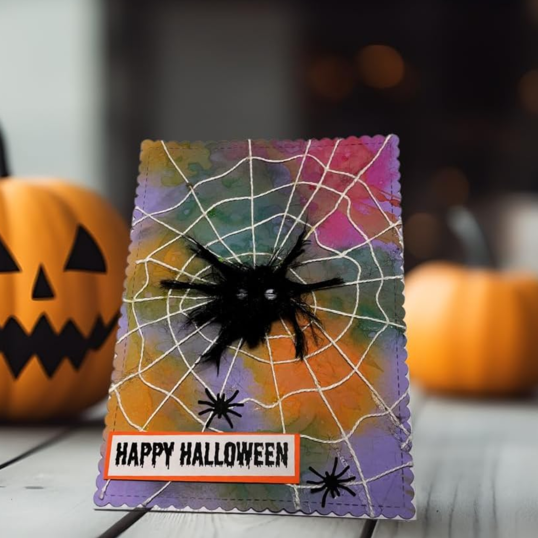 A handmade Halloween greeting card with a colorful watercolor background, white spiderwebs, a black feather spider, and a 'HAPPY HALLOWEEN' banner, displayed on a wooden surface with pumpkins in the background.