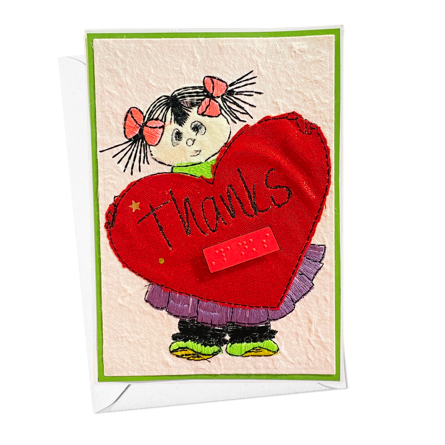 A applique stitched handmade-style greeting card featuring a cartoon girl with black pigtails and pink bows holding a large red heart that says 'thanks' in black script. The card has a green border, a textured cream background, and is shown with a white envelope behind it on a white background