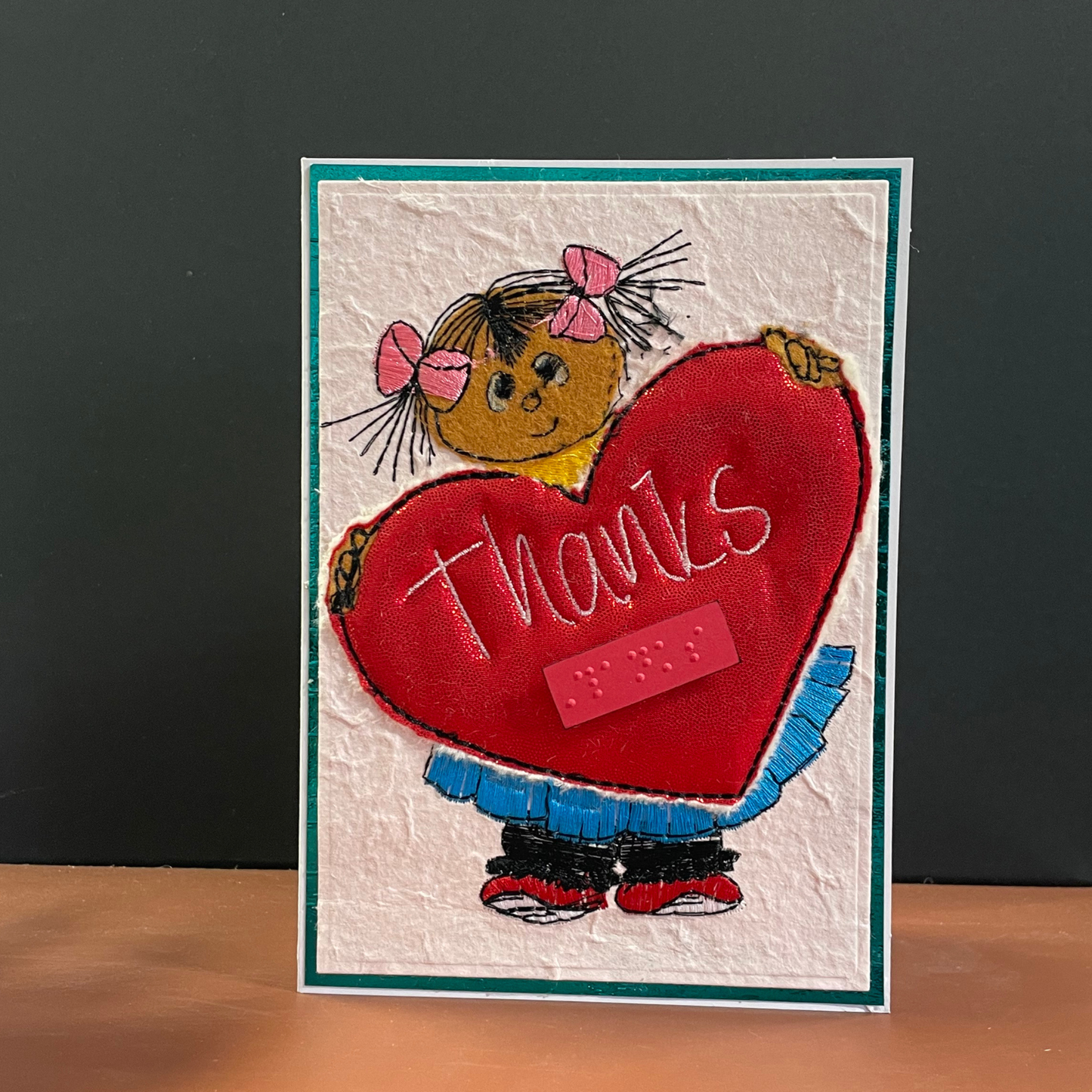 A handmade greeting card featuring a cartoon girl with pink bows holding a large red heart that says 'thanks' in white script, set against a textured off-white background with a teal border.