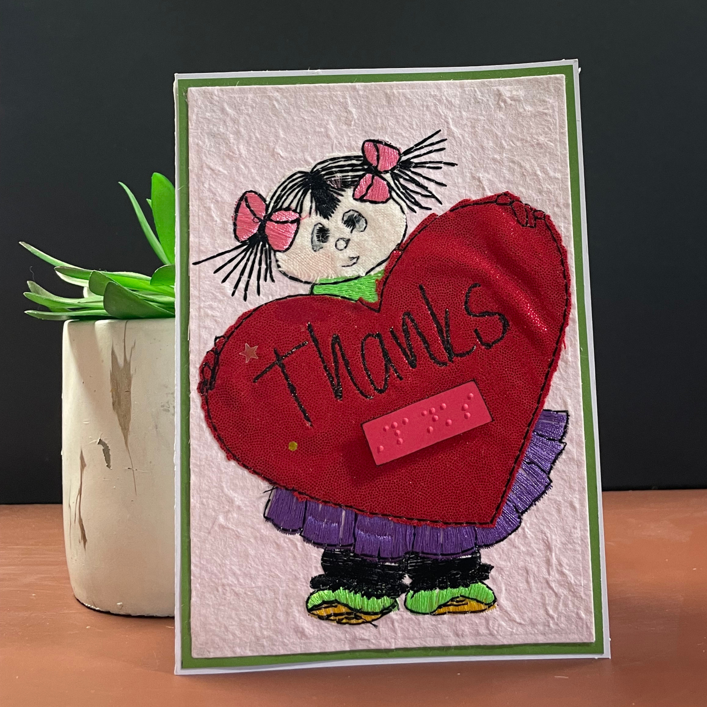 A handmade  card with a green border, featuring a cartoon girl with black pigtails and pink bows holding a large red heart that says 'thanks' in black script. The card is displayed upright on a wooden surface next to a small potted succulent against a dark gray background.