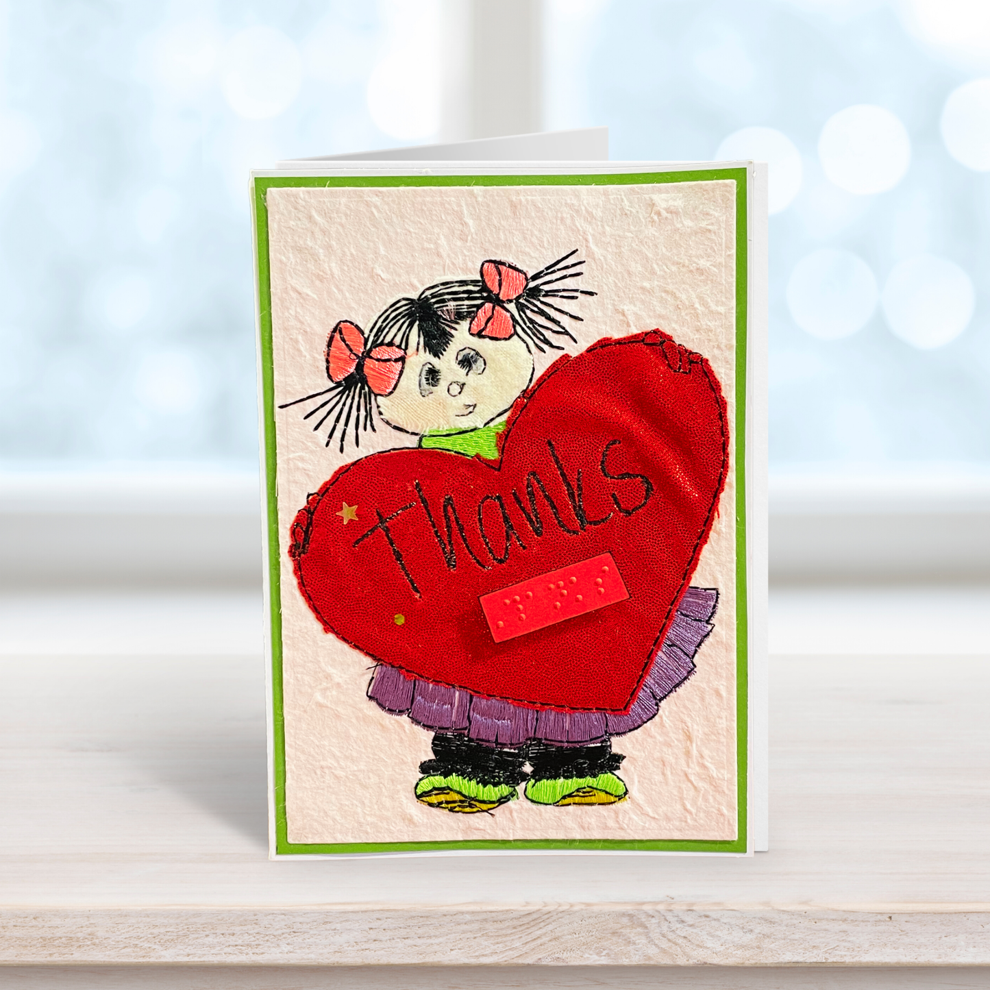 A handmade greeting card featuring a cartoon girl with black pigtails and pink bows holding a large red heart that says 'Thanks' in script, standing on a light wooden surface with a soft, blurred bokeh background.