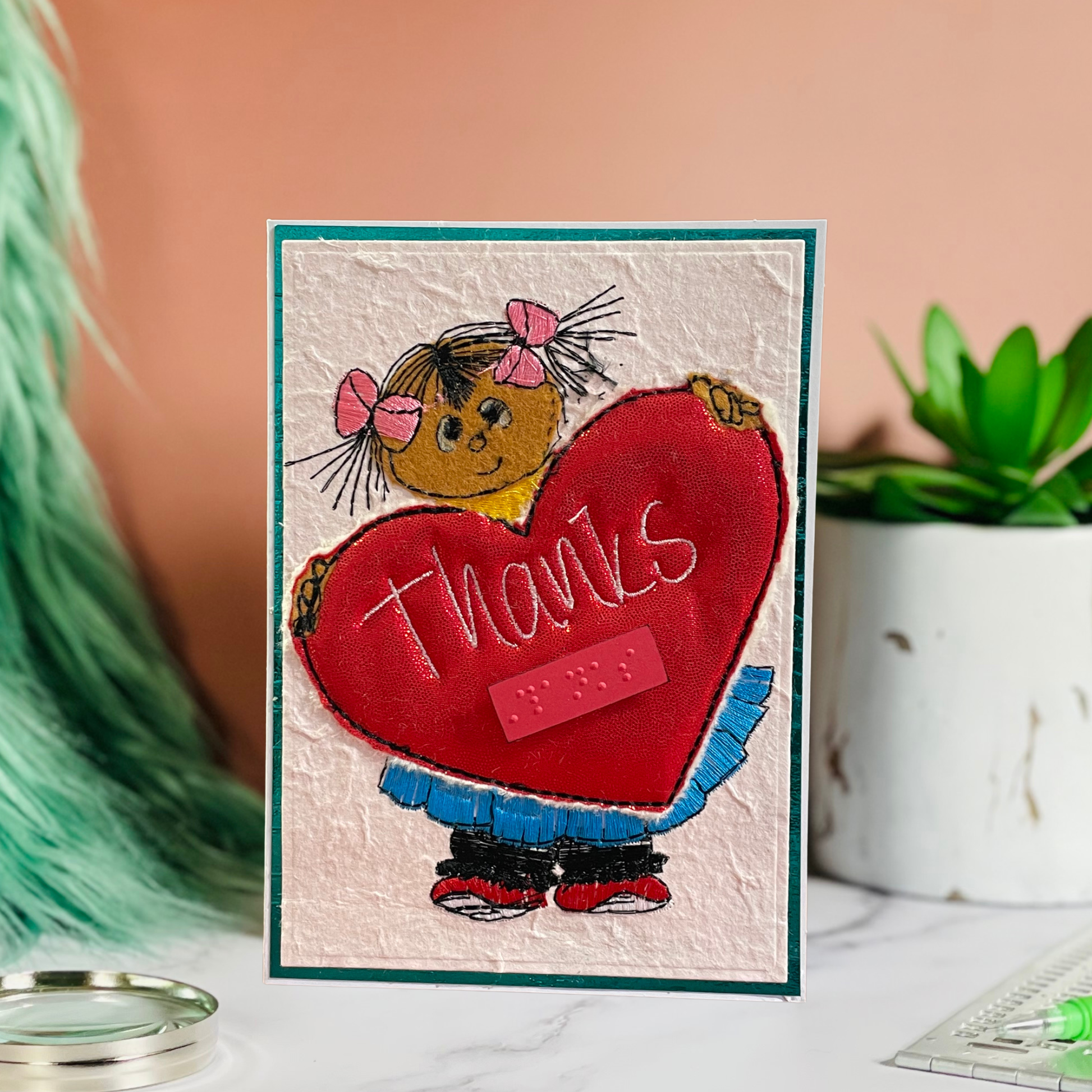A handmade greeting card featuring a cartoon girl with pink bows holding a large red heart that says 'thanks' in white script, styled on a marble surface with a peach background and green plant.