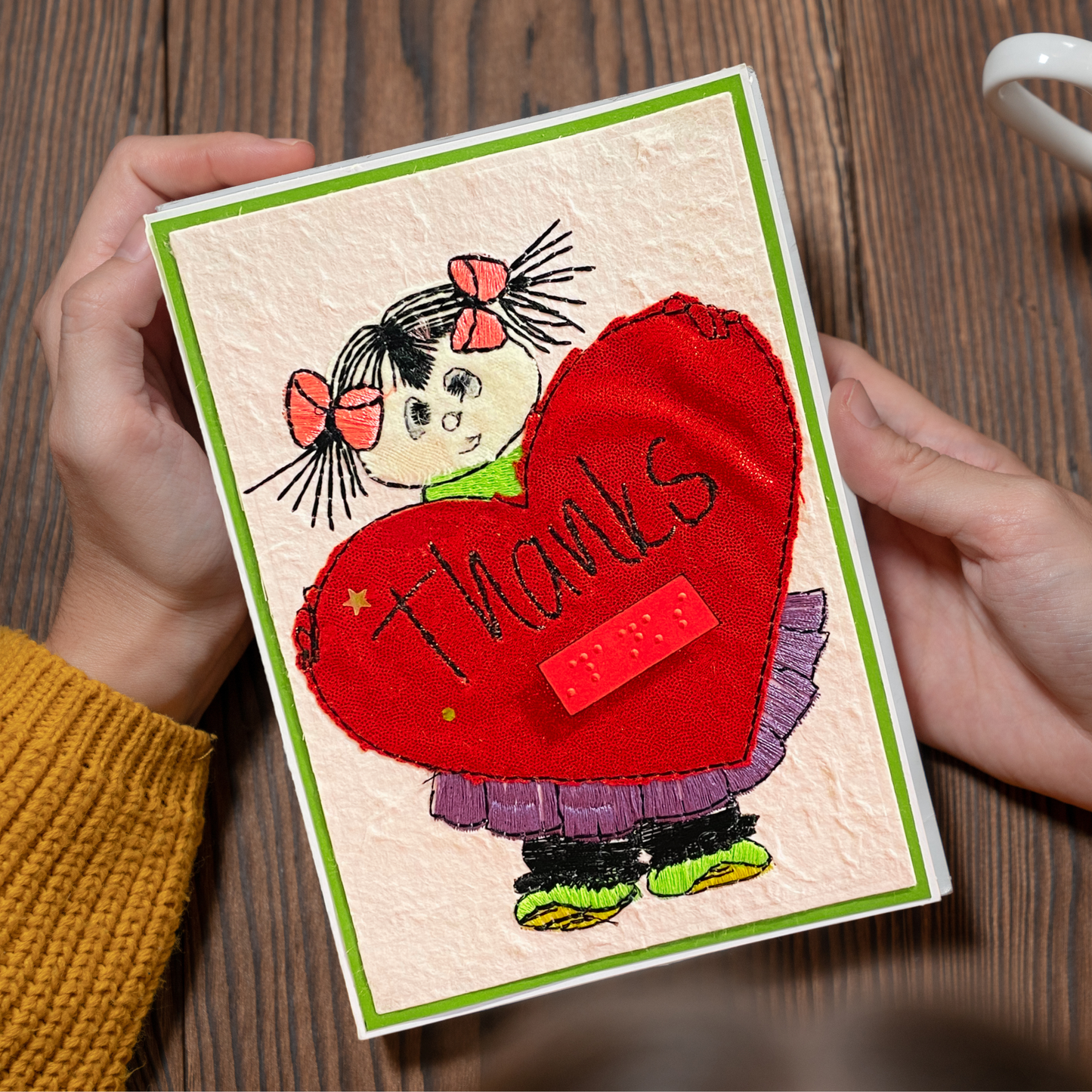 A handmade textured card held by two hands over a wooden table. The card features a cartoon girl with pink bows holding a large red heart that says 'thanks' in black script, with a green border.