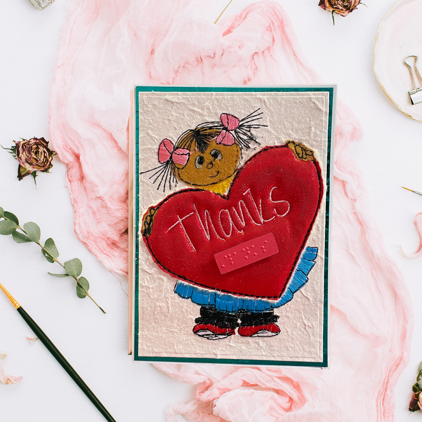 A handmade greeting card featuring a child holding a large red heart with the word 'thanks' on it, styled on a white surface with pink fabric, dried flowers, and a paintbrush.