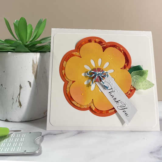 A handmade layered flower greeting card with orange and yellow petals, a small blue and white ribbon bow, and a 'Thank You' tag, displayed on a marble surface next to a potted succulent.