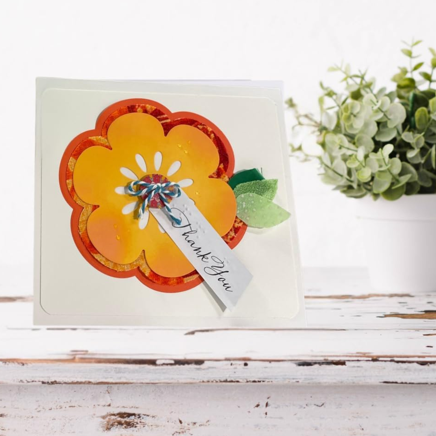 A handmade layered flower card with orange and yellow petals, green leaves, and a small 'Thank You' tag, displayed on a rustic white wooden surface with a blurred green plant in the background.