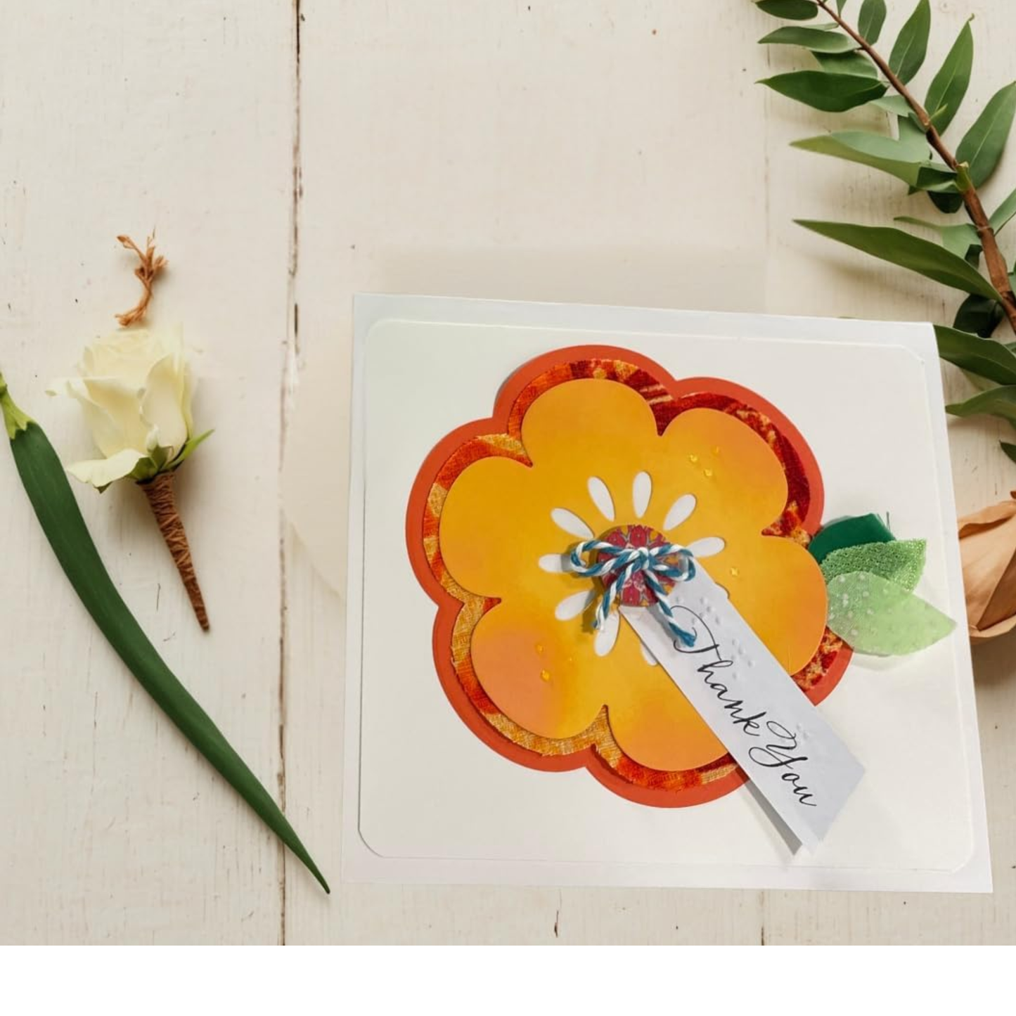 A handmade layered flower greeting card with orange and yellow petals, a small blue and red bow, and a 'Thank You' tag, displayed on a white wood background with decorative green leaves and a white rose.