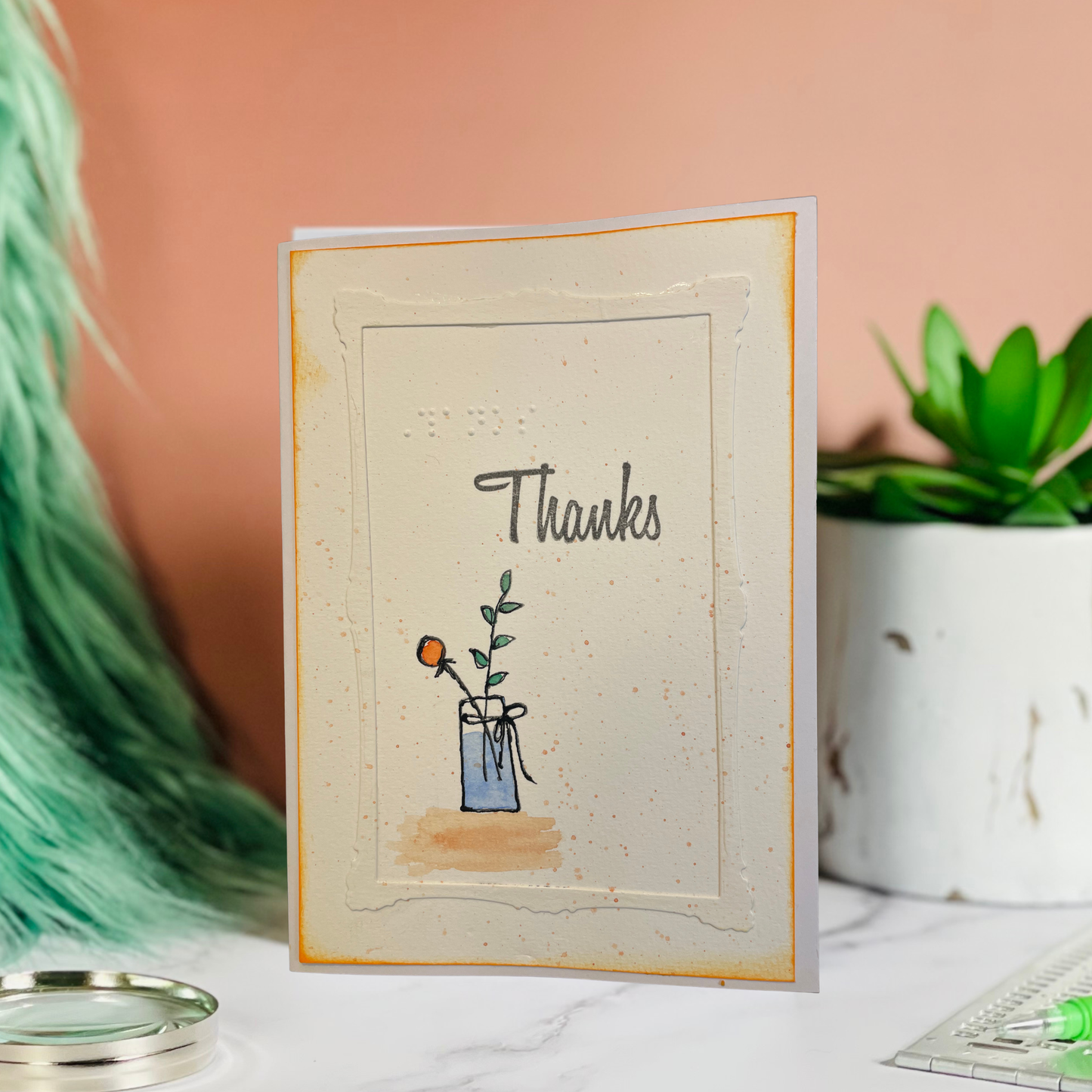 A handmade greeting card with 'Thanks' written in script above a small blue vase illustration, standing on a marble surface with a peach background and decorative plants.
