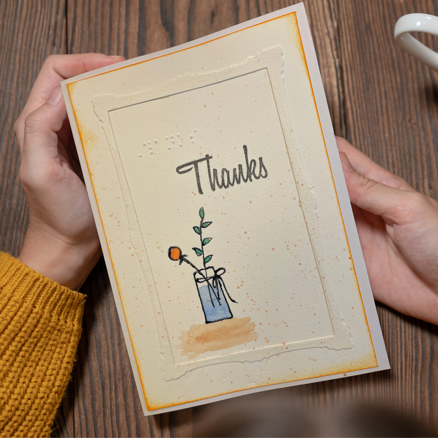 A handmade square greeting card held by two hands over a wooden table. The card has a cream background with a raised border, the word 'Thanks' in gray script, and a small illustration of a blue vase with a green stem and orange flower.