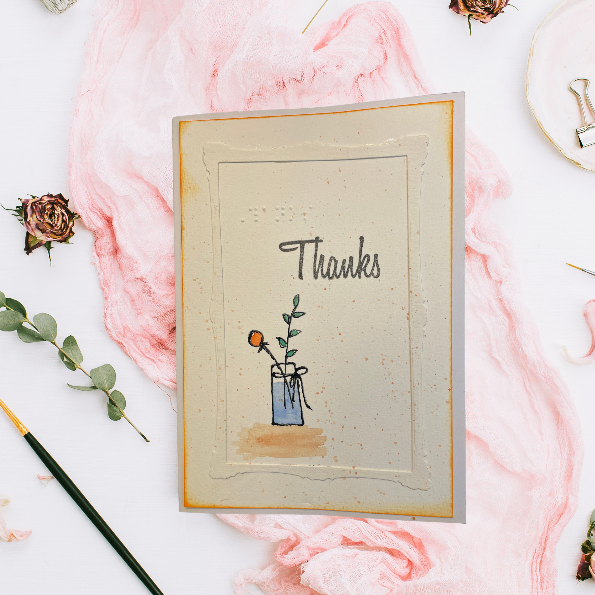 A handmade thank you card with a textured cream background, featuring a small illustration of a flower in a blue vase and the word 'Thanks' in script. The card is styled in a flat-lay arrangement with pink paper, dried roses, a paintbrush, and eucalyptus on a white surface.