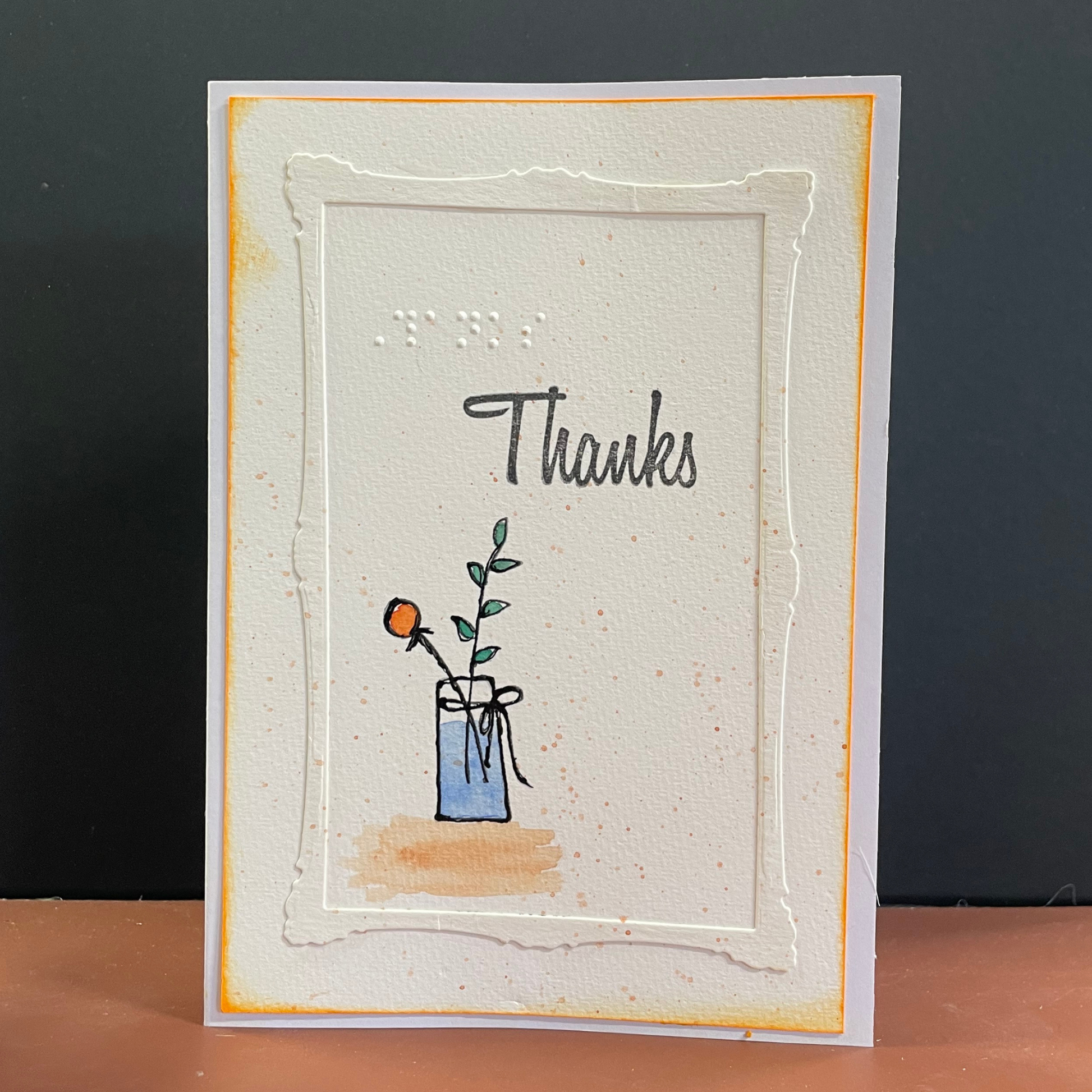 A handmade layered card with a white embossed frame, yellow-orange border accents, and the word 'Thanks' in black script above a small illustration of a flower in a blue vase, set against a dark gray background.