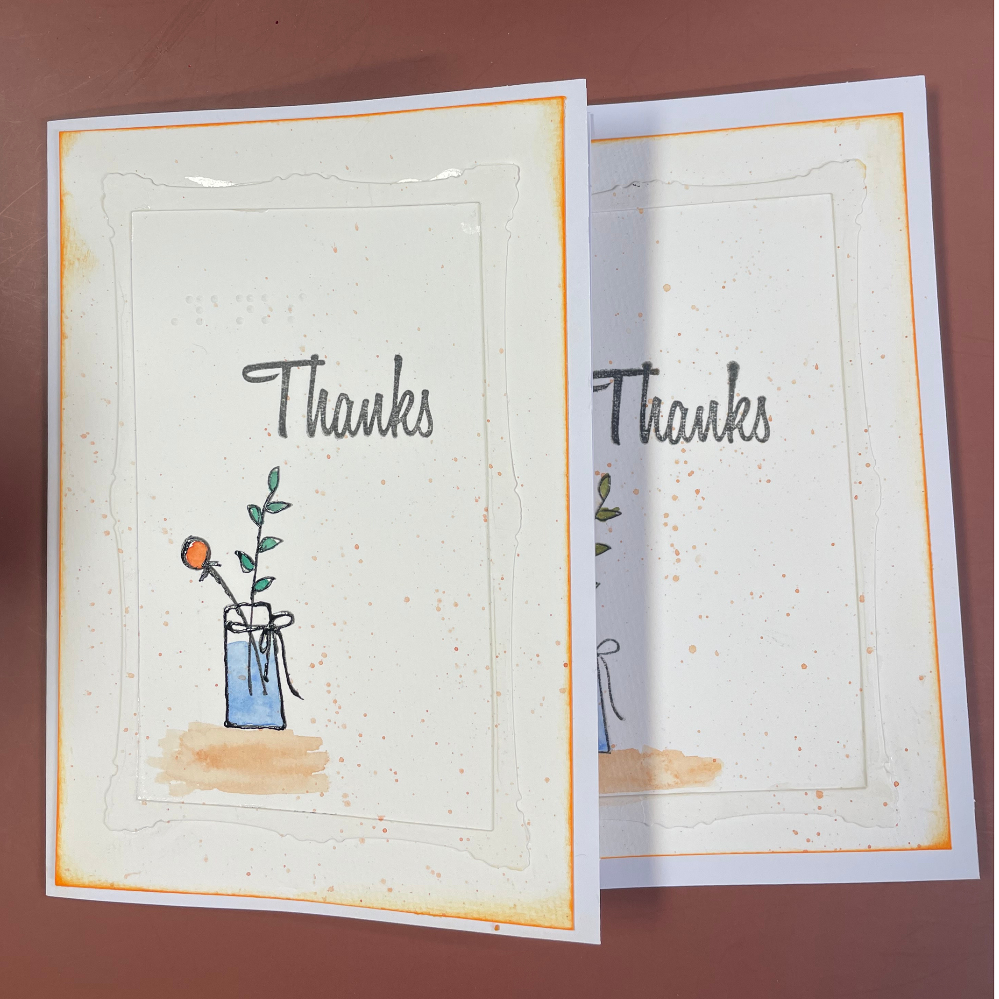 Two handmade greeting cards with a watercolor design of a flower in a blue vase and the word 'Thanks' written in script, displayed on a brown background.
