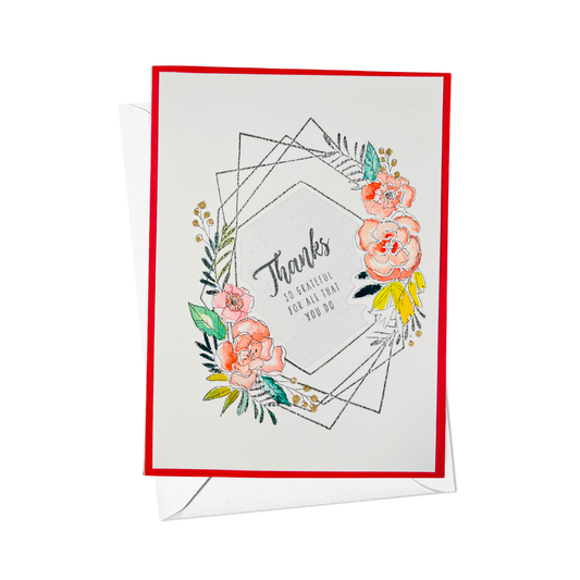A greeting card with a red border, featuring a floral wreath design in pink, green, and yellow around the word 'Thanks' and the phrase so greatful for all you do.' in text and braille. A white envelope is partially visible behind the card on a clean white background.