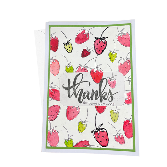 A greeting card with a white background, featuring hand stamped strawberries in pink, red and green, with a central white label that reads 'thanks for thinking sweet' in print and braille.