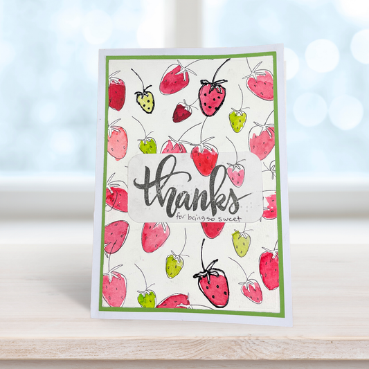 A handmade greeting card featuring a white background decorated with watercolor-style red and pink strawberries, green stems, and a green border. The center has a clear window over black script reading 'thanks for baking a sweet' with small text underneath.
