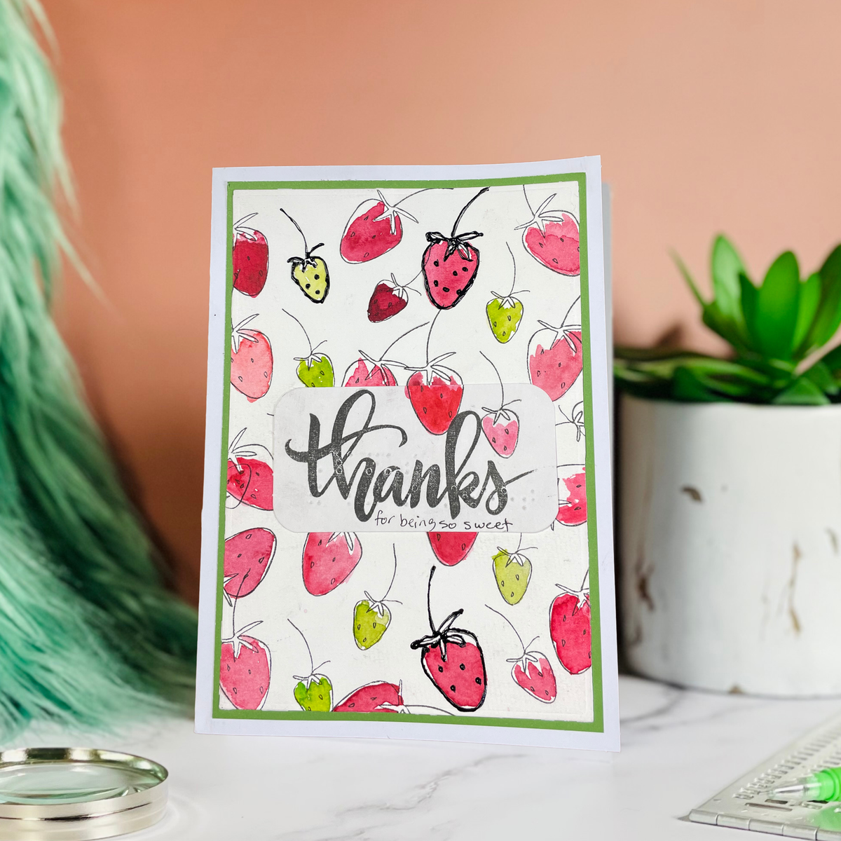 A handmade greeting card with a white background, green border, and illustrations of red and green strawberries, featuring the text 'thanks for baking sweet' in black script. The card is standing on a marble surface with a peach-colored wall, a potted succulent, and other decorative items in the background.