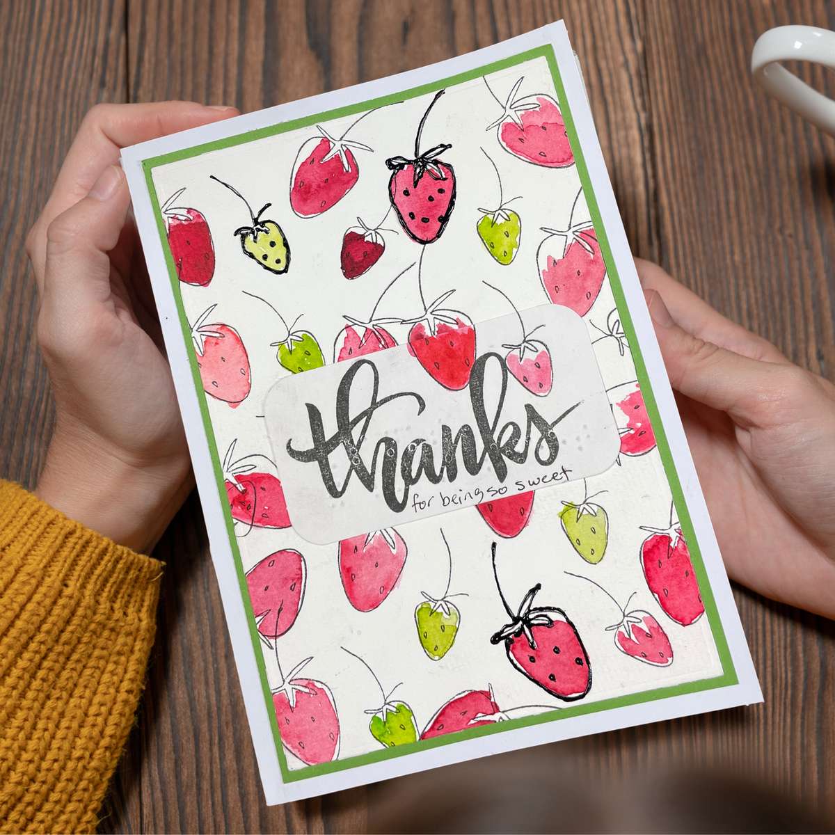 A handmade greeting card with a strawberry pattern and the word 'thanks' in script, held by two hands over a wooden table. The card has a green border, a white base, and illustrations of red and green strawberries.