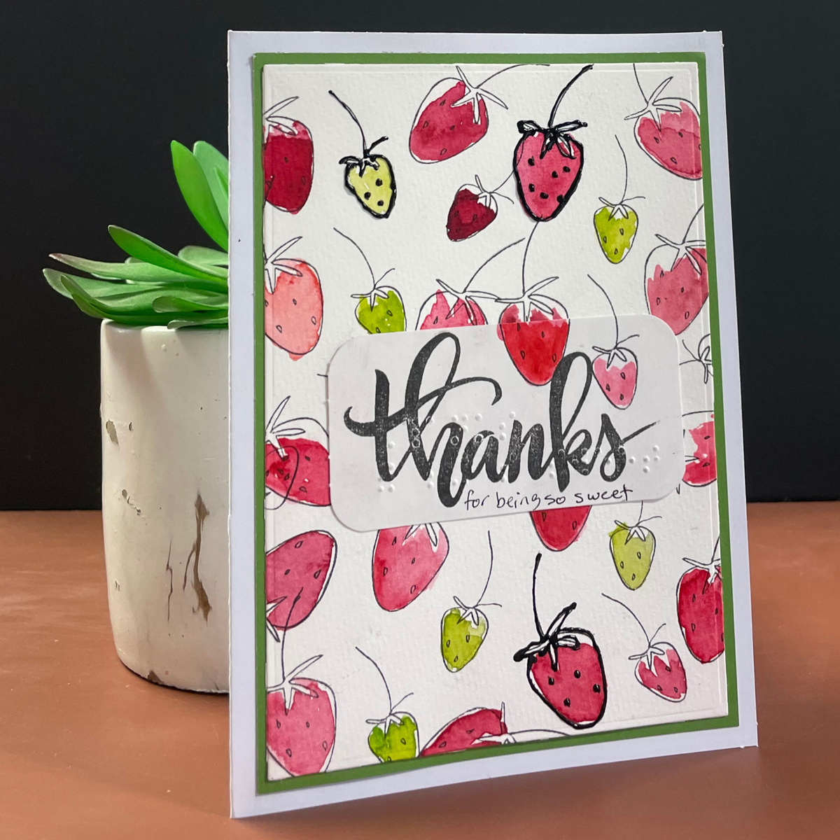 A handmade greeting card with a white background, decorated with a pattern of red and green hand-stamped strawberries. A central white banner features the cursive text 'thanks for bringing so sweet' in print and braille
