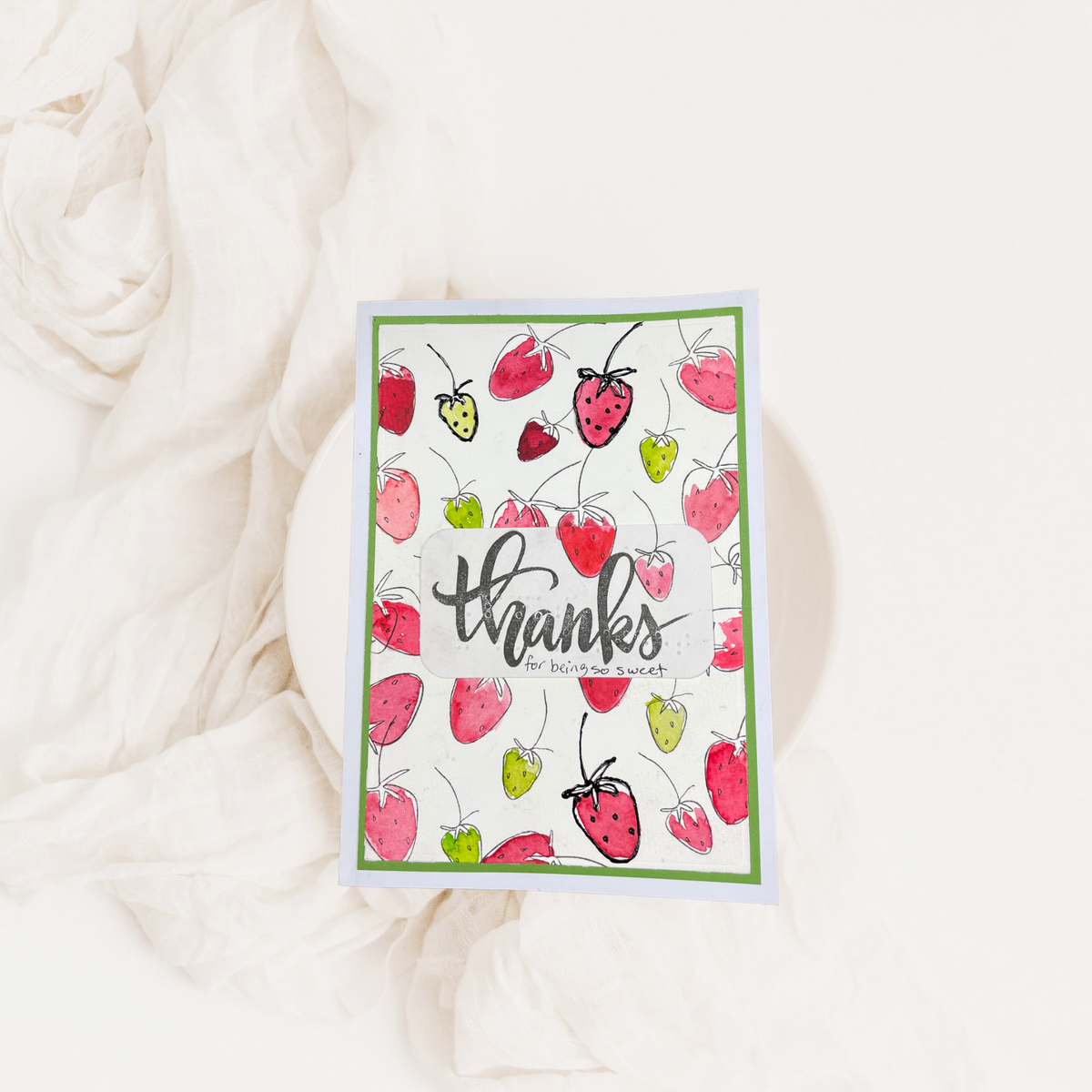 A handmade card featuring a watercolor strawberry pattern and the word 'thanks' in black script, styled on a white plate with draped white fabric.