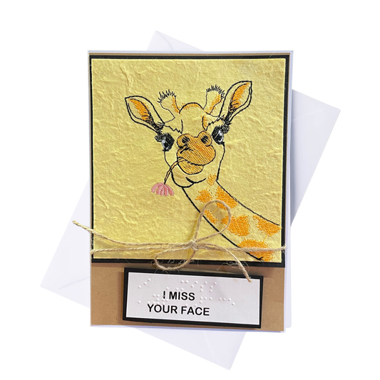 A handmade greeting card featuring a textured yellow background with an embroidered giraffe holding a pink flower, mounted on a brown kraft paper with a jute twine bow and a white label that reads 'I MISS YOUR FACE' with braille. A white envelope is visible behind the card on a white background.