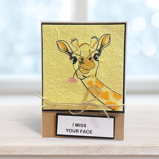 A handmade greeting card featuring a textured yellow background with an embroidered giraffe illustration and a kraft brown base. A small white label at the bottom reads 'I MISS YOUR FACE' in black text, and the card is tied with natural twine.
