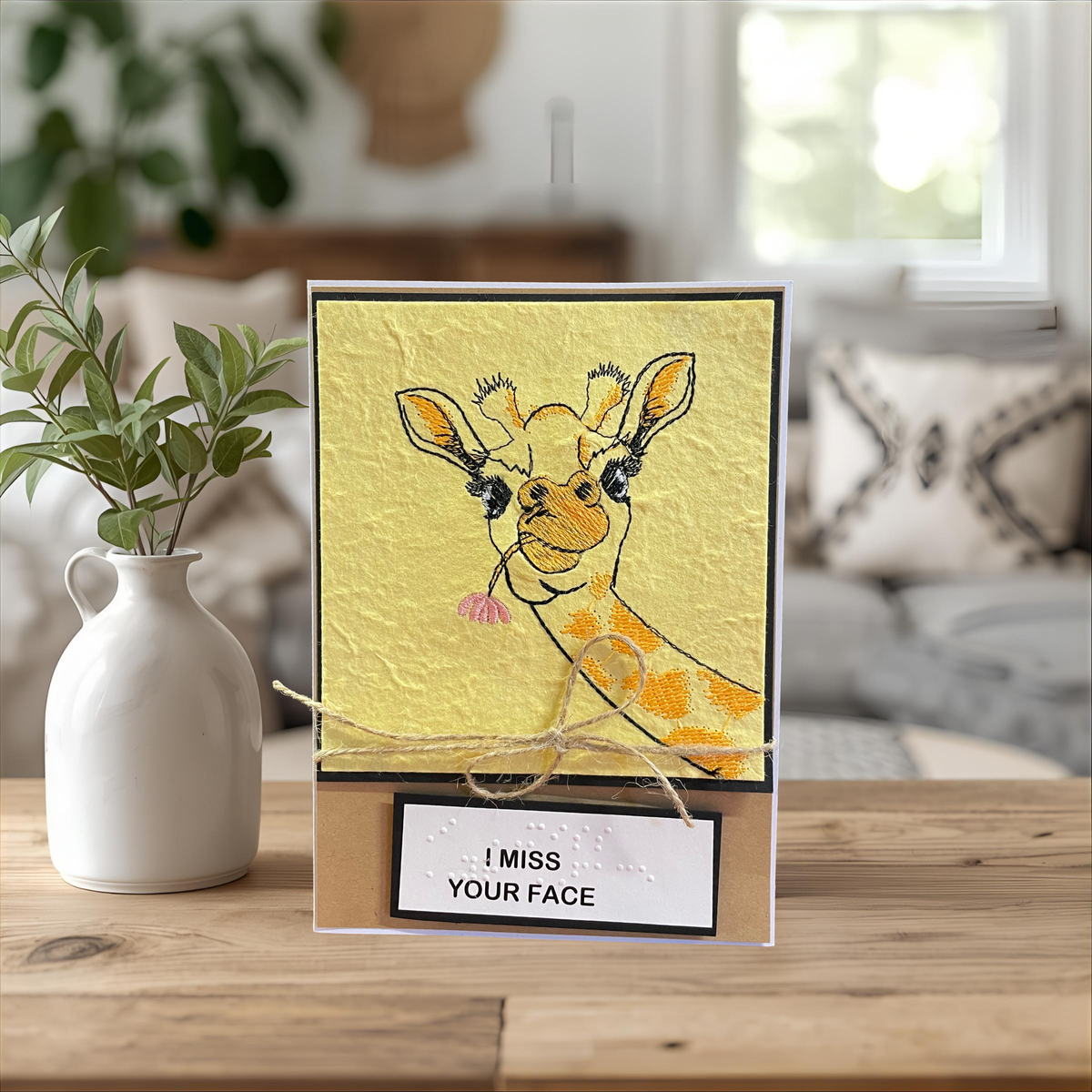A handmade greeting card featuring a textured yellow background with an embroidered giraffe holding a pink flower, tied with twine and resting on a wooden surface next to a white ceramic vase with green leaves. The card has a wooden base with a white label that reads 'I MISS YOUR FACE'.