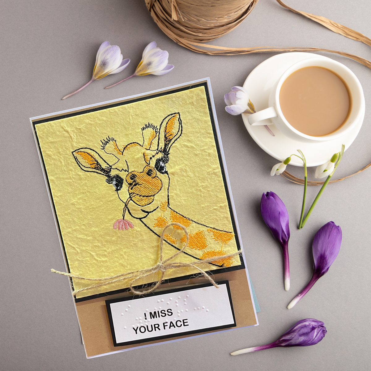 A handmade greeting card featuring a textured yellow background and an embroidered of a giraffe with the text 'I MISS YOUR FACE' in Braille. The card is styled in a flat-lay arrangement on a gray surface alongside a cup of tea, twine, and purple flowers.
