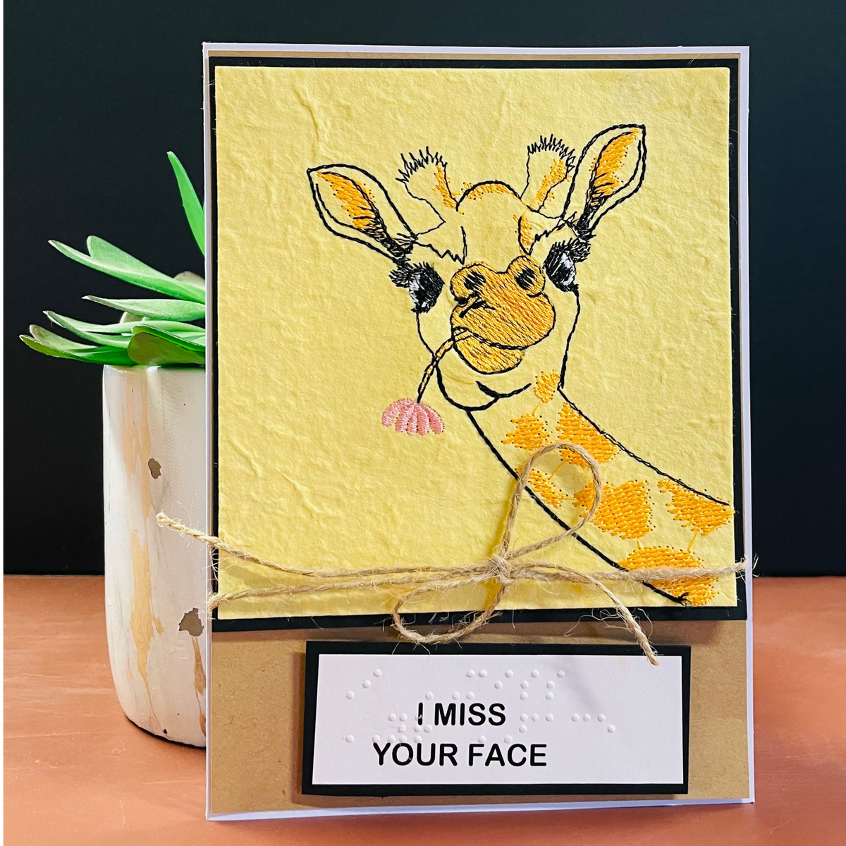 A handmade greeting card featuring a textured yellow background with an  embroidered of a giraffe eating a pink flower. The card is decorated with a rustic twine bow and a white label at the bottom that reads 'I MISS YOUR FACE' in print and braille