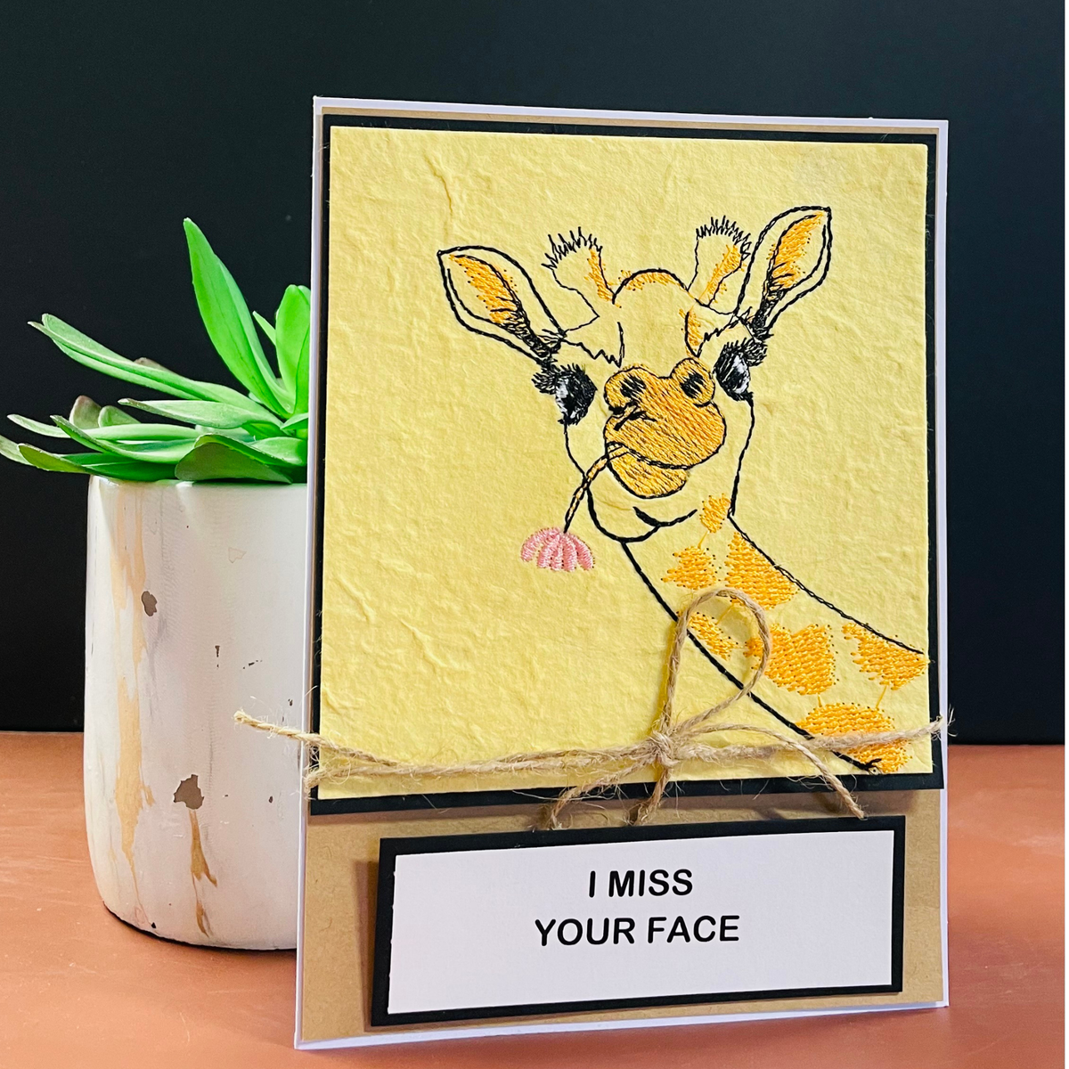 A handmade greeting card featuring a textured yellow background with an  embroidered of a giraffe eating a pink flower. The card is decorated with a rustic twine bow and a white label at the bottom that reads 'I MISS YOUR FACE'.