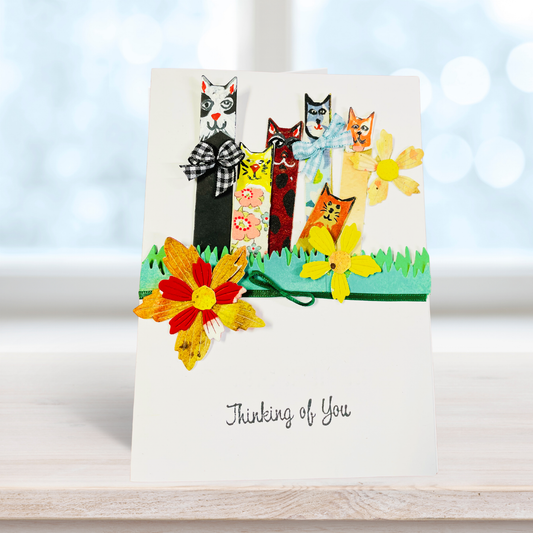A handmade greeting card featuring five whimsical, colorful cats standing on a green grassy strip, with layered paper flowers in front and the text 'Thinking of You' printed at the bottom. The card is displayed standing upright on a light wooden surface against a soft, blurred white background with bokeh light.