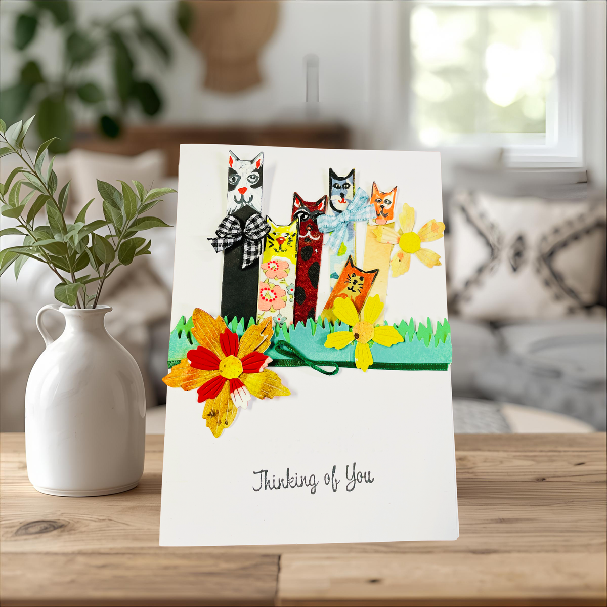 A handmade 'Thinking of You' greeting card featuring four whimsical cat figures in autumn colors, displayed on a wooden table next to a white ceramic vase with green leaves. The background shows a softly blurred living room interior.