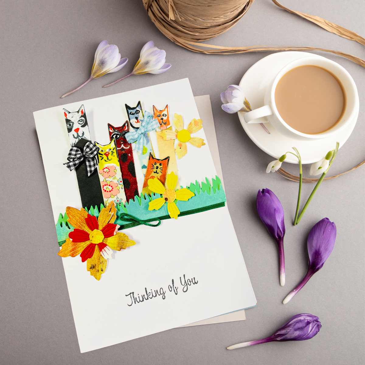 A handmade greeting card featuring colorful paper cats and flowers with the text 'Thinking of You', styled in a flat-lay arrangement with a cup of coffee, purple flowers, and twine on a gray background.