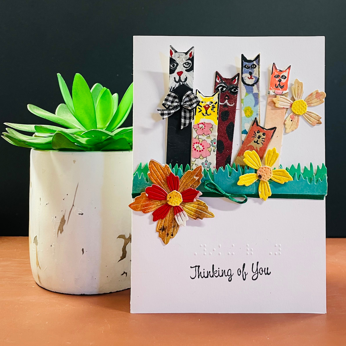 A handmade greeting card with a white background, featuring four whimsical stick-figure cats in different colored outfits standing behind green grass, decorated with colorful paper flowers. The card has 'Thinking of You' written in black script at the bottom.