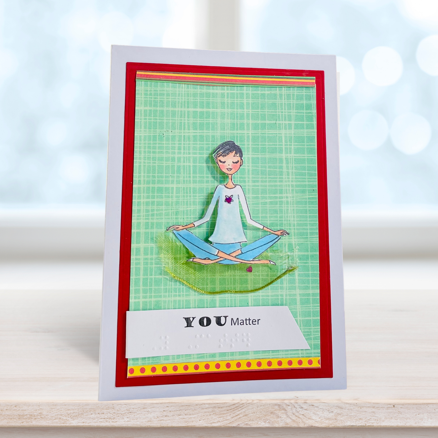 A handmade greeting card featuring a cartoon illustration of a woman meditating on a green grid background, framed with red and yellow borders. The bottom reads 'YOU Matter' with Braille text.