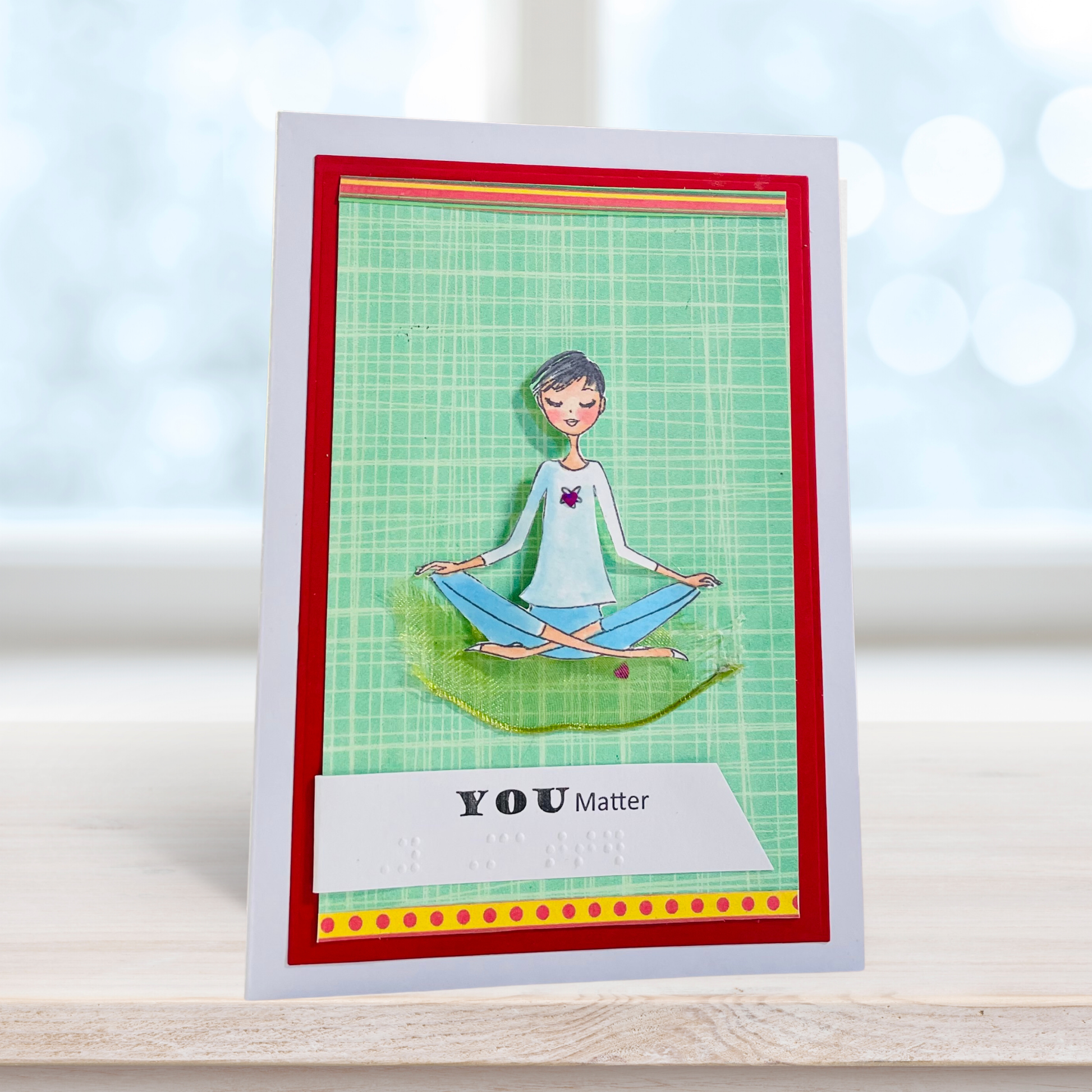 A handmade greeting card featuring a cartoon illustration of a woman meditating on a green grid background, framed with red and yellow borders. The bottom reads 'YOU Matter' with Braille text.