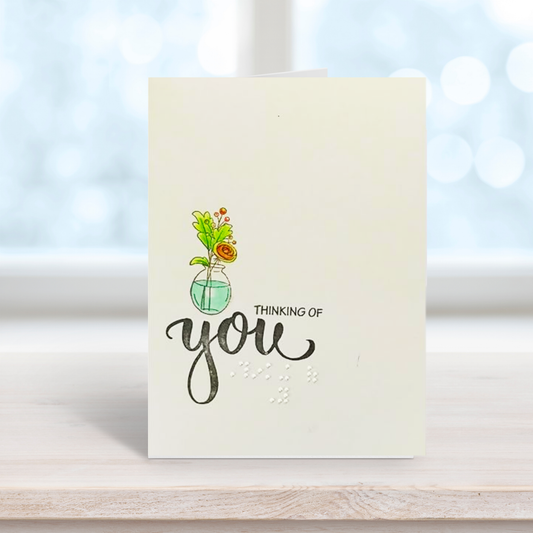 A greeting card with a watercolor illustration of flowers in a glass vase and the words 'THINKING OF you' in black script and braille 