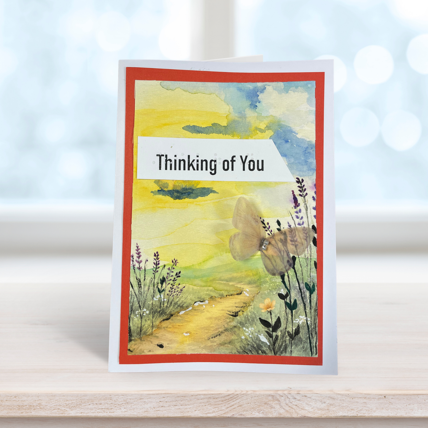 A handmade greeting card standing upright on a light wooden surface. The card features a watercolor landscape with a yellow and green field, a butterfly, wildflowers, and a red border.