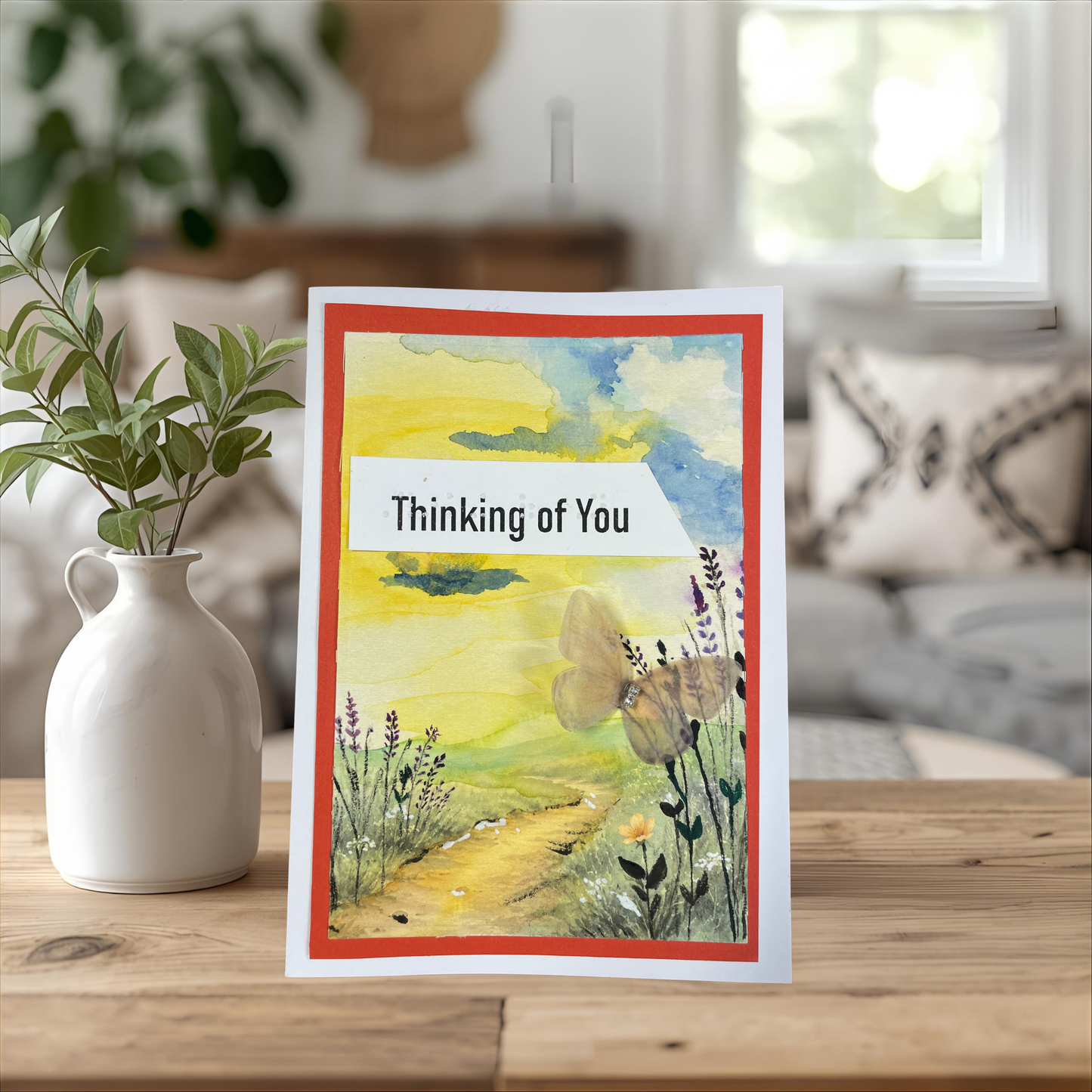 A greeting card with a watercolor landscape illustration and the text 'Thinking of You' in a white banner, standing on a wooden table next to a white ceramic vase with green leaves. The background is a softly blurred living room interior.